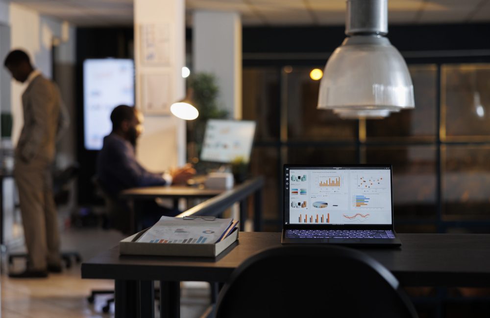 Laptop computer with marketing statistics standing on desk table in startup office, businessmen working overtime at company strategy. African american entrepreneurs brainstorming business ideas